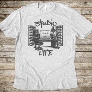May include: A light gray t-shirt with a black graphic of a recording studio setup. The design includes a desk with a computer, speakers, a microphone, and the words "Studio Life".
