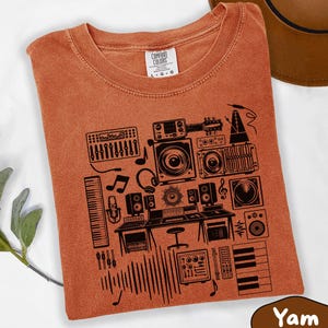 May include: A rust-colored t-shirt featuring a black graphic print of various music production equipment, including a mixing console, speakers, a keyboard, a microphone, headphones, and musical notes, with the Comfort Colors brand label at the neck.