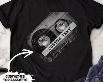 May include: A black t-shirt featuring a graphic of a vintage cassette tape positioned at an angle. The cassette tape has a white label area with the black text CUSTOM TEXT printed on it. The design has a distressed, textured appearance.
