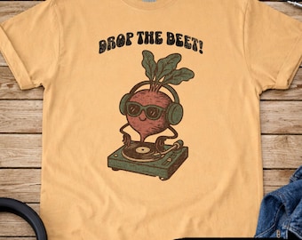 May include: Mustard-colored t-shirt with the text "DROP THE BEET!" above a cartoon beet wearing headphones and sunglasses, playing a record on a turntable. The word "MUSTARD" is at the bottom.