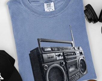 May include: A light blue t-shirt featuring a black and white graphic of a boombox. A black baseball cap and headphones are also visible. The shirt has the text "Blue Jean" on the bottom right.