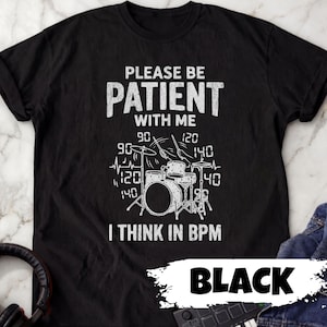 May include: A black short-sleeved t-shirt featuring a white graphic of a drum kit with drumsticks and the text "PLEASE BE PATIENT WITH ME I THINK IN BPM" surrounded by various numbers including 90, 120, and 140, along with a heartbeat line.