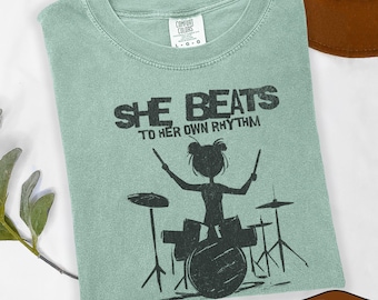 May include: A light green Comfort Colors t-shirt with the text "SHE BEATS TO HER OWN RHYTHM" and a black silhouette of a drummer playing a drum set. The shirt is a size L-G-G.
