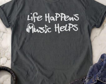 May include: A dark gray short-sleeved t-shirt featuring the white text "Life Happens Music Helps" with a graphic of headphones replacing the letter "u" in the word "Music". The shirt has a crew neckline and a standard fit.