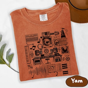 May include: A burnt orange t-shirt featuring a black graphic of a music studio setup, including speakers, a mixing board, and musical notes. The shirt has a crew neck and short sleeves. The tag reads "Comfort Colors".