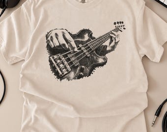 May include: A beige short-sleeved t-shirt featuring a black graphic print of a bass guitar being played. The design shows a detailed illustration of hands positioned on the fretboard and body of the instrument, rendered in a stippled, ink-like style.