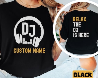 May include: Black t-shirt with a white DJ logo featuring headphones and sound waves. The back of the shirt says "Relax the DJ is here" in white text.  The shirt is shown on a model wearing blue jeans.