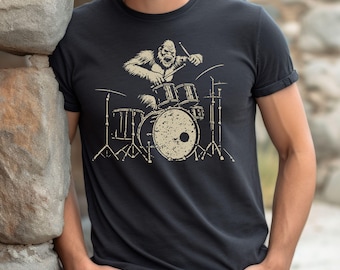 May include: Black t-shirt featuring a beige illustration of a gorilla playing a drum set. The gorilla is depicted with its mouth open, and the drum set is detailed with various drums and cymbals. The shirt has short sleeves and a crew neck.