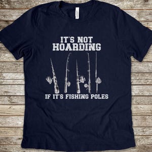 May include: Navy blue t-shirt with a white graphic that says "It's not hoarding if it's fishing poles". The graphic also includes a row of four fishing poles with reels.