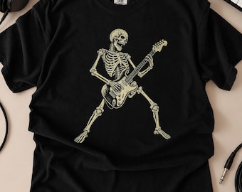 May include: A black short-sleeve t-shirt featuring a graphic print of a skeleton playing an electric guitar. The skeleton is depicted in a dynamic pose, rendered in a light beige color. The shirt has a crew neckline and a Comfort Colors brand label.