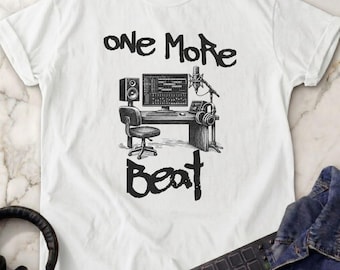 May include: A white t-shirt featuring a black ink-style illustration of a music production studio setup with a computer, speaker, microphone, and chair. The text "one More Beat" is printed in a stylized, hand-drawn font above and below the graphic.