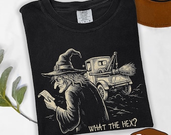 May include: A black t-shirt featuring a graphic illustration of a witch in a pointed hat holding a parking ticket, standing next to a tow truck with a broom attached to the back. The text "WHAT THE HEX?" is printed in white at the bottom.