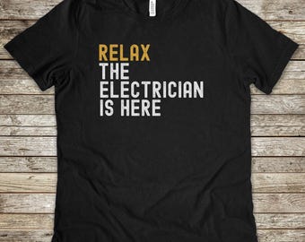 May include: A black short-sleeved t-shirt featuring the text RELAX in gold lettering, followed by THE ELECTRICIAN IS HERE in white lettering. The design has a distressed, weathered texture and is printed on the front of the crew neck garment.
