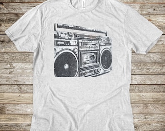May include: A light gray t-shirt featuring a black and white graphic of a vintage boombox. The boombox design includes speakers, a cassette player, and the text "NEW YORK". The shirt has a crew neck and short sleeves.