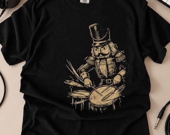 May include: A black short-sleeve t-shirt featuring a graphic print of a nutcracker soldier playing a drum set. The illustration is rendered in a muted gold tone, depicting the soldier in a uniform with a tall hat, playing drums and cymbals.