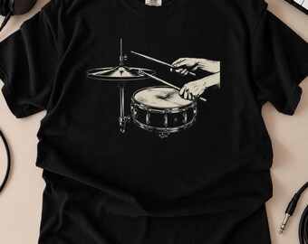 May include: A black short-sleeve t-shirt featuring a cream-colored graphic print of a snare drum and a cymbal on a stand. The illustration depicts two hands holding drumsticks, with one stick positioned over the cymbal and the other over the drum.