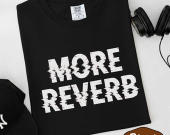 May include: Black t-shirt with the text "MORE REVERB" in white, featuring a glitch effect. A black baseball cap and headphones are also visible. The shirt is labeled "Comfort Colors".