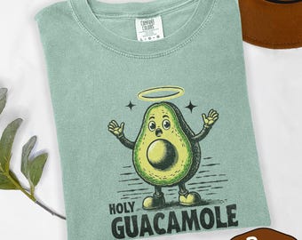 May include: A light green t-shirt with a cartoon avocado character wearing a halo and the words "Holy Guacamole". The avocado has arms and legs and is smiling. The shirt is made of a soft material.