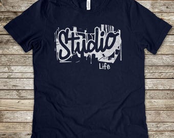 May include: Navy blue t-shirt with the words "Studio Life" in a white, graffiti-style design. The text is layered over a stylized cityscape. The shirt is made of a soft material.