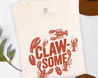 May include: Ivory-colored t-shirt with a red lobster design and the word "CLAW-SOME". The shirt is made of a soft material and is a comfortable fit. The shirt is a great gift for anyone who loves lobsters.
