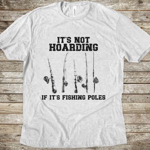 Op de afbeelding: Een lichtgrijs t-shirt met korte mouwen en een zwarte afbeelding van vijf hengels. De tekst boven de hengels luidt IT'S NOT HOARDING en de tekst onder de hengels luidt IF IT'S FISHING POLES in een vetgedrukt, verweerd zwart lettertype.