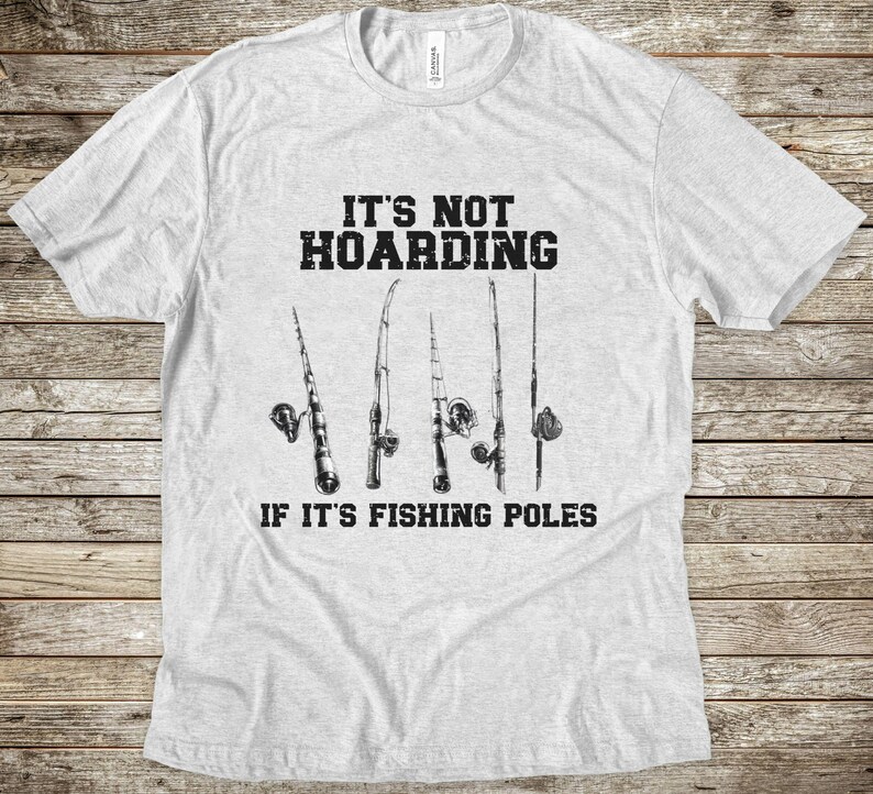 Op de afbeelding: Een lichtgrijs t-shirt met korte mouwen en een zwarte afbeelding van vijf hengels. De tekst boven de hengels luidt IT'S NOT HOARDING en de tekst onder de hengels luidt IF IT'S FISHING POLES in een vetgedrukt, verweerd zwart lettertype.