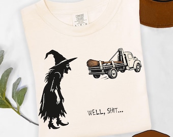 May include: An ivory t-shirt featuring a black graphic of a witch standing next to a tow truck carrying a broomstick. The text "WELL, SHIT..." is printed in black below the truck. The shirt has a Comfort Colors brand label at the neckline.