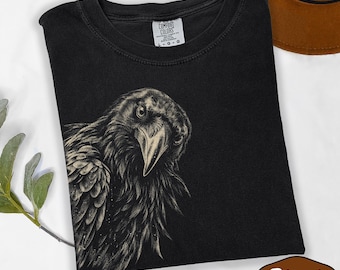 May include: A black short-sleeve t-shirt featuring a detailed graphic illustration of a raven in shades of cream and grey. The shirt has a crew neckline and a Comfort Colors brand label printed on the inside collar with the size L clearly visible.