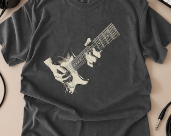 May include: A charcoal gray short-sleeve t-shirt featuring a graphic print of a guitar being played. The design shows a detailed illustration of hands positioned on the fretboard and body of an electric guitar, rendered in a muted off-white tone.