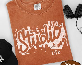 May include: A yam-colored t-shirt with the word "Studio" in a white graffiti-style font. The word "Life" is printed below. A black baseball cap and headphones are also visible.