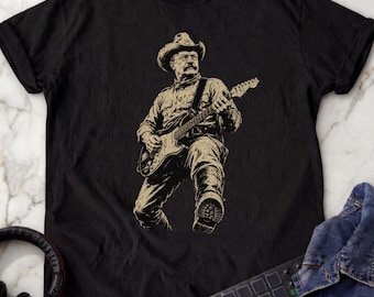 May include: A black short-sleeved t-shirt featuring a beige graphic print of a person wearing a cowboy hat and boots while playing an electric guitar. The illustration shows the figure in a dynamic pose with one leg raised, rendered in a detailed sketch style.