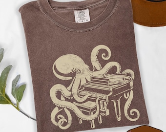 May include: A brown short-sleeve t-shirt featuring a cream-colored graphic of an octopus playing a grand piano. The shirt has a crew neckline and a Comfort Colors brand label on the inside collar. The text on the label reads Comfort Colors L G G.