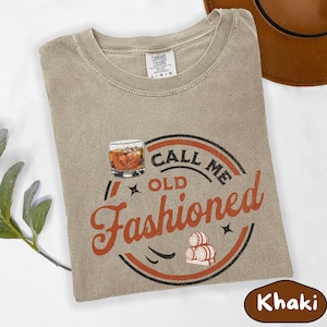 May include: Khaki-colored t-shirt with a crew neck and the words "Call Me Old Fashioned" in a circular design. The design includes a whiskey glass, barrels, and stars. The shirt is made of a soft material.