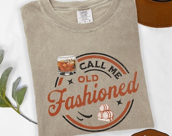 May include: Khaki-colored t-shirt with a crew neck and the words "Call Me Old Fashioned" in a circular design. The design includes a whiskey glass, barrels, and stars. The shirt is made of a soft material.