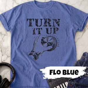 May include: A blue t-shirt with the words "TURN IT UP" in distressed black lettering. Below the text is a black and white illustration of a hand turning a dial. The shirt is labeled "Flo Blue".