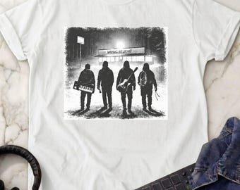 May include: A white t-shirt featuring a black and white graphic of four figures standing before a building labeled MUSIC STUDIO. The figures carry musical instruments including a keyboard and a guitar, set against a dark, textured background.