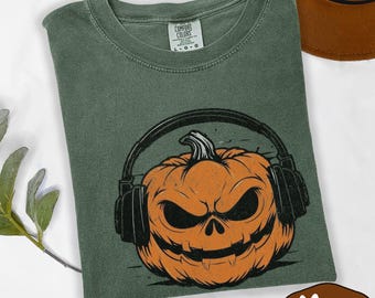 May include: A moss green t-shirt featuring a jack-o'-lantern wearing headphones. The pumpkin has an angry expression and is orange with black details. The shirt has the Comfort Colors logo in the upper center.
