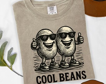 May include: A khaki crew neck t-shirt featuring a graphic of two anthropomorphic beans wearing sunglasses and giving a thumbs up. The text "COOL BEANS" is printed in black below the illustration. The shirt has a Comfort Colors brand label.
