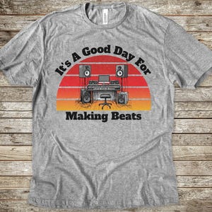 May include: A heather gray t-shirt featuring a graphic of a music production studio with speakers, a keyboard, and a computer. The design is set against a red and orange sunset background with the text It's A Good Day For Making Beats.