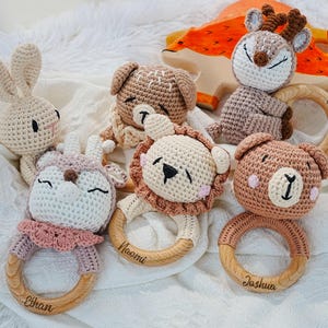 May include: A selection of handmade crochet baby rattles shaped like animals, such as a bunny, dog, deer, lion, and bear. Each rattle has a wooden ring and is in soft, neutral colours.