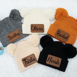 Può includere: Cinque berretti di lana con pon pon, ognuno con un toppa di pelle personalizzata. I berretti sono di vari colori, tra cui grigio, bianco, arancione, beige e nero. Le toppe hanno i nomi Bartley, Olivia, Rachel, Trista e Noah.