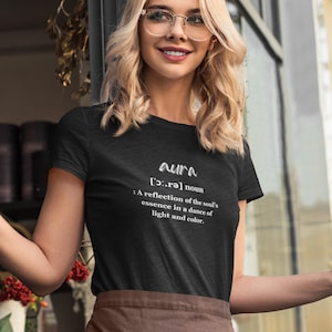 May include: Black t-shirt with the word "aura" and its phonetic pronunciation, along with a definition: "A reflection of the soul's essence in a dance of light and color." The shirt is worn by a person with blonde hair and glasses.