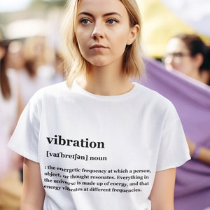 May include: A white t-shirt with the word "vibration" and its definition printed in black. The definition explains that everything in the universe is made up of energy and vibrates at different frequencies.