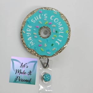 May include: A blue and gold donut-shaped badge holder with the text "Maybe She's Complete" and colorful sprinkles. The badge holder is attached to a clear plastic retractable badge reel with a blue glitter button.