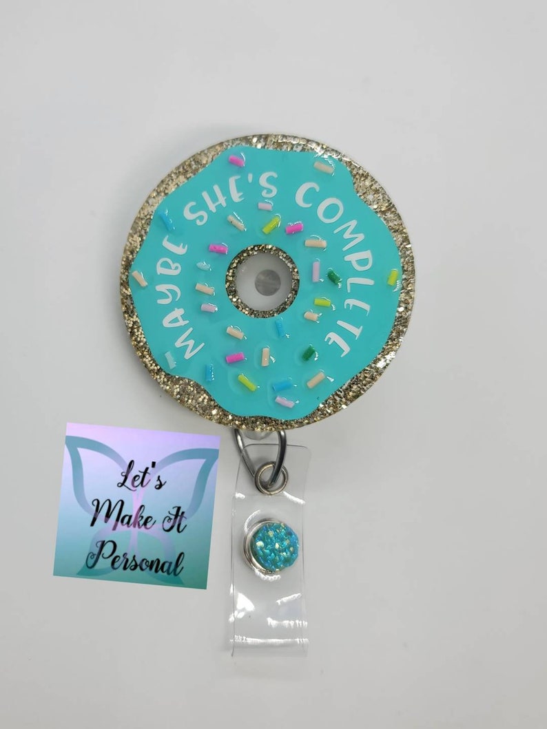 May include: A blue and gold donut-shaped badge holder with the text "Maybe She's Complete" and colorful sprinkles. The badge holder is attached to a clear plastic retractable badge reel with a blue glitter button.
