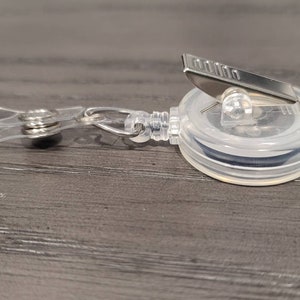 May include: A clear plastic retractable badge holder with a silver clip. The holder has a black retractable cord.