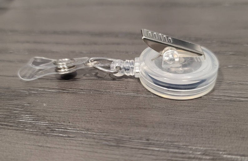 May include: A clear plastic retractable badge holder with a silver clip. The holder has a black retractable cord.