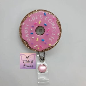 May include: Pink donut-shaped badge holder with gold glitter and sprinkles. The donut has the text "Maybe She's Complete" written around it.