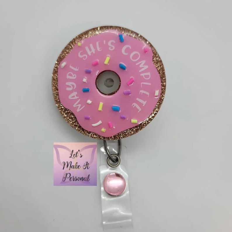 May include: Pink donut-shaped badge holder with gold glitter and sprinkles. The donut has the text "Maybe She's Complete" written around it.