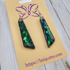 May include: A pair of dangle earrings with a black and green glitter resin design. The earrings are displayed on a card with a butterfly design and the text "https://1mip.etsy.com".